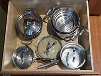 Stainless steel pots and pans with lids inside a drawer, including a steamer insert and frying pan.