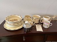 Top-view image showing 2 bowls, 1 plate, 3 Bunnykins fine bone china cups, 1 ABC baby cup, and 1 metal baby sippy cup on wooden surface