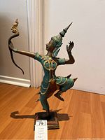 Side angle view of a green, gold, and red painted metal statuette depicting a traditionally dressed Siamese figure in a dynamic pose, holding a curled ornamental element.