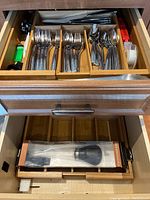 Drawer with organized stainless steel flatware including forks, spoons, knives, and wooden trays holding the pieces. Also shows dripless baster in original packaging.