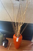 Front view of a tall orange wood vase filled with pale willow branches on a wooden floor.