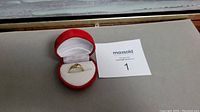 Ring displayed in red velvet box with white padded interior, placed on a surface next to a card with lot number