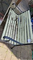 Outdoor heavy duty bench with peeling paint and metal curved armrests shown on paved surface.