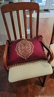 Photo of a wooden dining chair with light beige cushioned seat, vertical slat back, and a decorative burgundy pillow with ornate embroidery and fringe.