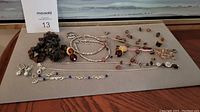 Photo of assorted costume jewelry pieces displayed on gray surface including necklaces, bracelets, and multiple earrings, mostly in silver-tone with some bead accents in dark red and black.