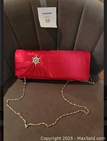 Front view of red satin evening bag with rhinestone starburst brooch and gold-tone chain strap on a brown chair.