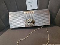 Silver evening clutch bag ribbed fabric with oval rhinestone brooch detail and silver chain strap, placed on grey cushioned seat.