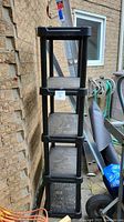 Stacked view of one black resin/plastic heavy duty adjustable shelving unit showing four shelves and vertical supports alongside a wall and ladder.