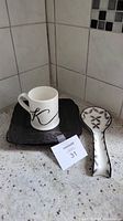 Photo showing Indigo mug, two black Mikasa plates beneath it, and decorative stoneware spoon rest with black and red pattern on countertop.