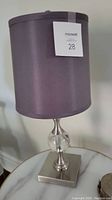Front view of the table lamp showing the gray fabric cylindrical shade, silver metal base with clear glass decorative element, placed on a marble-like surface.