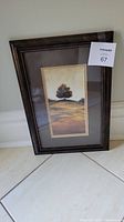 Framed landscape print leaning against a wall on tile floor, showing the full frame and matting.
