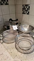 Photo showing stainless steel pressure cooker with lid and black handles, surrounded by three stainless steel colanders of varying sizes and types.