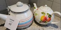 Two teapots on a kitchen counter, one porcelain with fruit design, one metal with stripes and white lid.