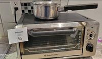 Toastmaster stainless steel toaster oven on countertop with small stainless steel saucepan on top. Oven has glass door with dial controls on right side, showing signs of use and needing cleaning.
