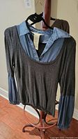 Grey knit sweater layered over blue satin-look long sleeve shirt on hanger, displayed on wooden hanger stand.