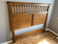 Queen size oak headboard with slatted top and panel design leaning against wall on wood floor, showing full front view.