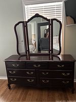 Front view of cherrywood triple dresser with ornately carved mirror frame, showing 9 drawers with brass handles, curved edges, and visible scratches on wood surface.