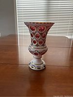 Full view of cranberry Bohemian glass vase showing flared top, decorative cutouts, hand-painted floral garlands and gold detailing on white overlay glass.