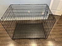 Front angled view of black metal dog crate with bottom tray on wooden floor.