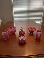 Full set of cranberry glass hobnail items displayed on table including two ruffled vases, cruet with stopper, candleholder, and two tumblers.