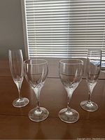 Four crystal glasses including two wine glasses and two champagne flutes arranged on a wooden table against window blinds background.
