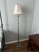 Full view of the tall floor lamp with brushed chrome metal shaft and ornate base, showing the lamp standing on wooden floor next to a wooden piece of furniture and iron bed frame.