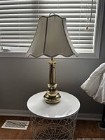 Full view of brass lamp with fitted cream fabric shade with scalloped edges, set on a white round table in front of a window with blinds.