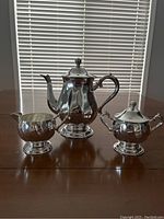 Silver tea set including teapot, creamer, and sugar bowl displayed together on a table in front of window blinds.