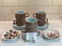 Overview of 6 stacked cups and saucers with 2 floral pattern side plates in front on white surface.