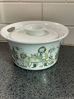 Side view of the covered casserole dish showing the Market pattern decoration featuring figurative characters in green and gold colors.
