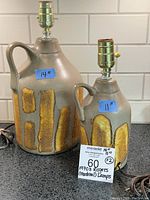 Photo showing both pottery jug lamps on a countertop with size labels 14" and 11" showing mustard yellow lava glaze detail on taupe pottery bases with lamp fittings.