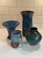 Four blue pottery pieces shown together on white counter with tiled wall background: raku vase, flared blue vase, ombre teal/black vase, small multicolor cup or vase.