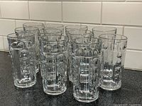 Eleven clear glass tumblers arranged on a kitchen countertop, showing alternating smooth and thumbprint dot panels in the vintage Heisey Old Sandwich pattern.