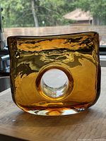 Front angled view showing the amber glass block vase with keyhole center, placed on light wooden surface with outdoor background visible.