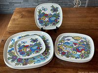 Set of 8 plates in colorful folk art Market Place pattern stacked on wooden table showing sizes and designs.
