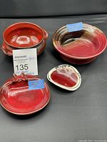 Four red hued pottery pieces shown on black background, including two bowls, one covered pot, and one small dish.