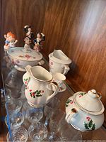 Wentworth China Christmas tea set items featuring creamer, sugar bowl with lid, small pitcher, teacup and saucer with holly and red berry decoration accented with gold trim, placed on glass shelf.