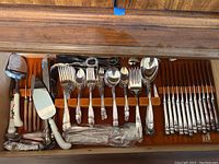 Overhead view of opened wooden flatware drawer with various knives, forks, spoons, and serving utensils arranged inside. White ceramic handled knives with red and green floral details, metal knives with tarnish, and forks and spoons with patterned handles.