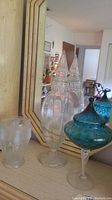 Three glass items on a surface including two tall and medium clear glass vases with pointed stoppers and a blue glass bowl with lid, all reflected in a mirror.