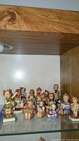 12 Goebel Hummel figurines arranged on a glass shelf inside a wooden cabinet showing various children figures in colorful outfits and playful poses.