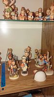 Multiple Goebel Hummel porcelain figurines of children and animals displayed on wooden shelves needing cleaning.