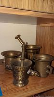 Four brass or metal mortars on wood shelf, three with pestles. Mortars vary in shape and size.