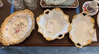 Three decorative serving platters displayed on a brown surface, showing two gold-rimmed floral platters and one ornate white hand-painted platter with gold accents.