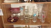 Nine assorted glass vases and bowls including rose- and plum-colored cut glass and clear glassware with Peter Pan text, arranged on a wood shelf.