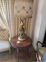 Full view of the vintage round wooden end table with carved details and the figurine lamp on top near seating furniture