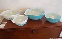 Four Pyrex mixing bowls displayed on wooden surface with numbered tags. Bowls include two large and two small sizes in white and blue with rooster and wheat designs.