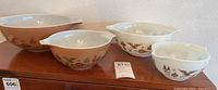 Four brown Pyrex mixing bowls on wood surface, varying sizes, white interiors, brown exteriors with vintage black motifs