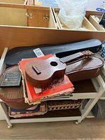 Ukulele resting on brown faux-snakeskin case with black hard case behind, atop a rolling cart