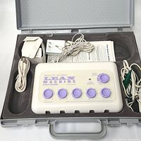 Open carrying case showing Beauty Works Lean Machine white and purple device with six knobs, power adapter, cables, and instruction sheet inside a gray case.