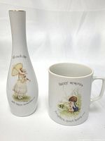 Photo of a white Holly Hobbie decorative vase with an illustration of a girl in a bonnet playing violin and text 'fill each day with happy memories' next to a mug with an illustration of a girl sitting outdoors and text 'happy memories freshen quiet hours'.